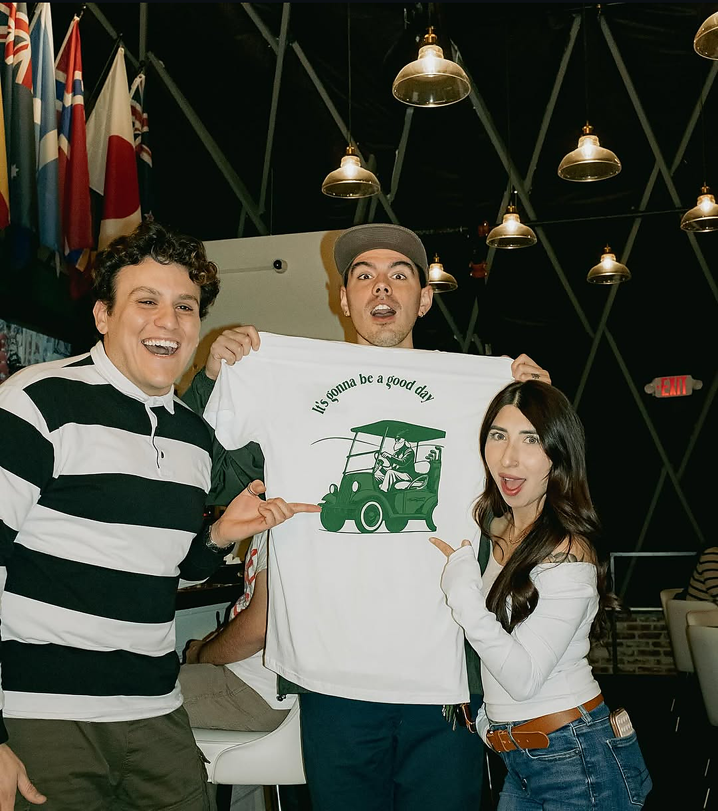 Three happy friends holding up white Mulligan's t-shirt reading It's gonna be a good day with golf cart illustration